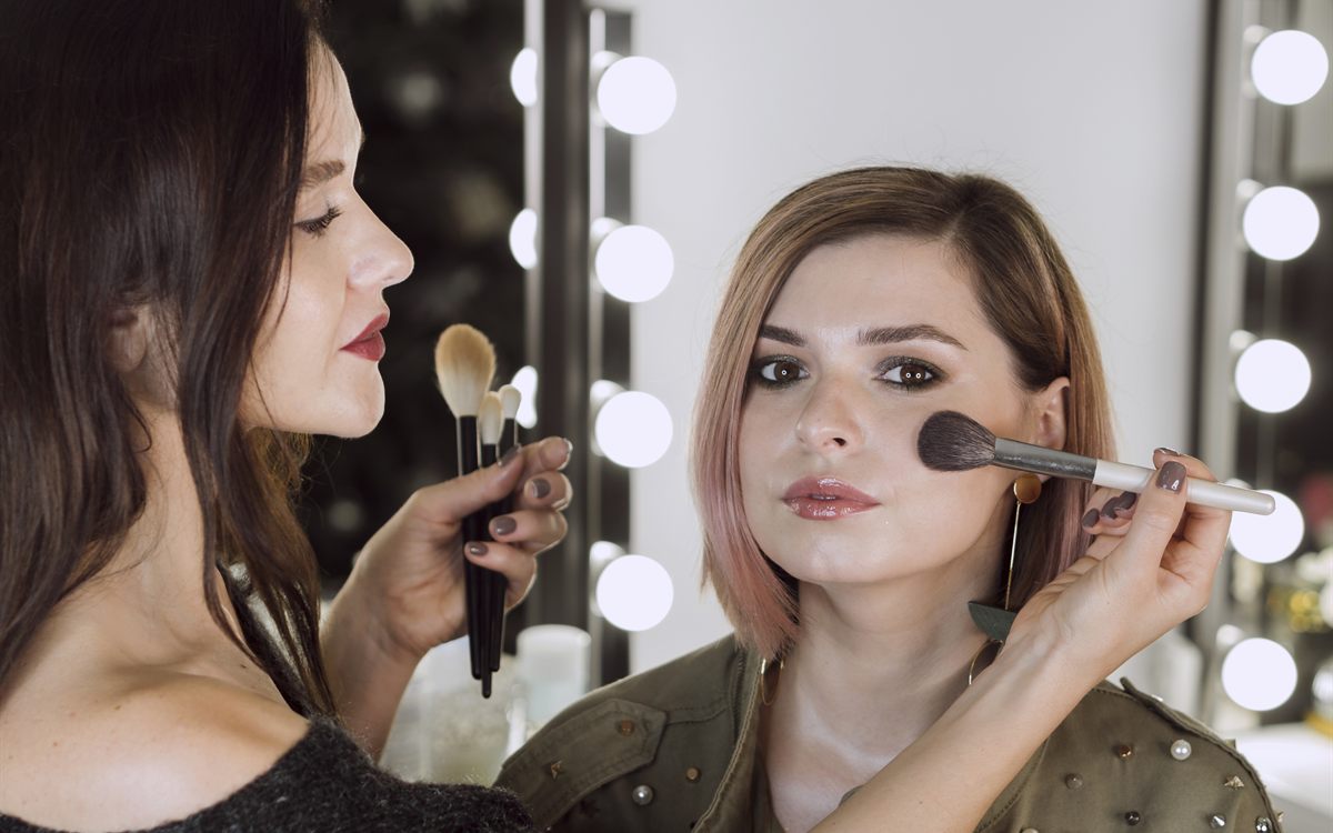 Makeup artist applying blush with a brush at a lit vanity mirror